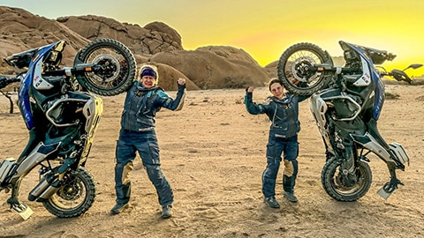 The riders on the German women’s team standing next to their motorcycles at sunset, with only the rear wheels of the motorcycles touching the ground. They each have a hand on the front wheel. The other hand is raised in a fist.